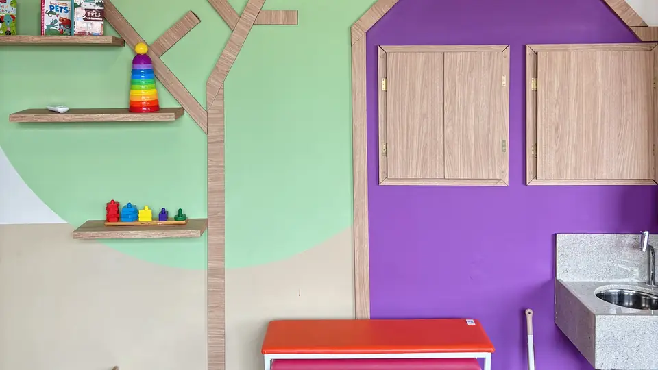 Sala de atendimento com parede verde, árvore de madeira com prateleiras e casinha lilás